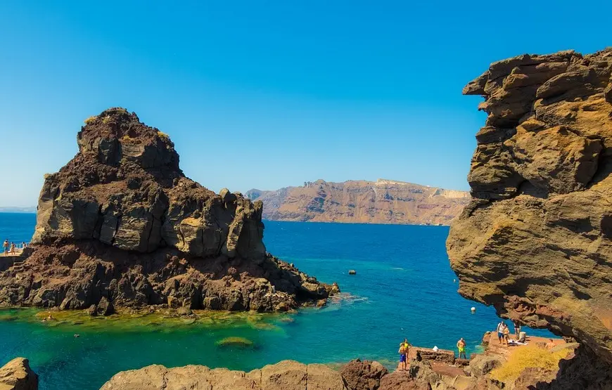 tra-le-rocce-rosse-di-santorini-si-affaccia-thirassia-isola-piu-piccola-il-mare-turchese-verde-intorno