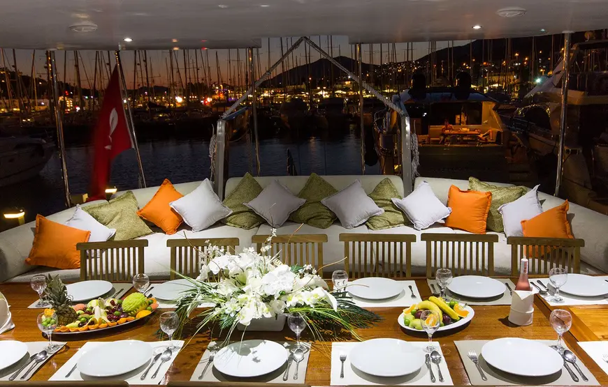 TAVOLATA-CON-PIATTI-BIANCHI-TOVAGLIETTE-UN-SOFA-CON-TANTI-CUSCINI-BIANCHI-ARANCIO-E-VERDI-CHE-GUARDANO-IL-PORTO-ILLUMINATO-CON-BARCHE-ORMEGGIATE