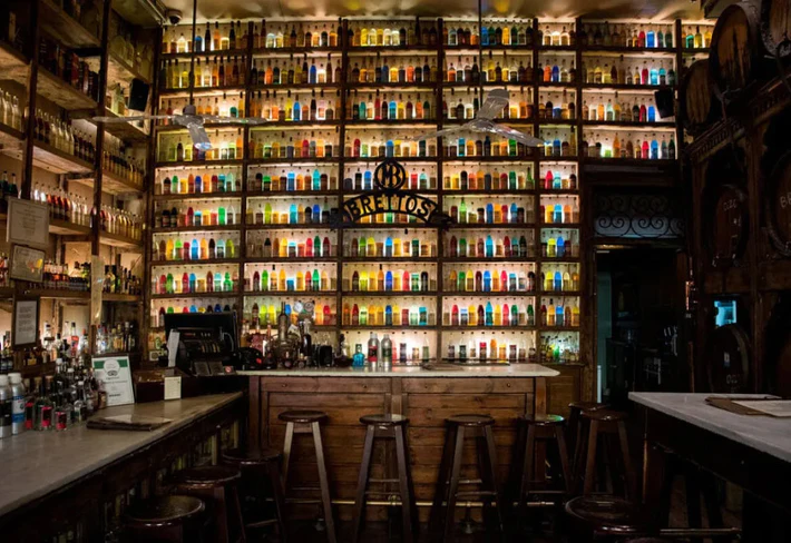 brettos-bar-con-parete-completamente-rivestita-di-bottiglie-colorate-retroilluminazione-la-scritta-brettos-al-centro-un-bancone-di-legno-atene-particolare