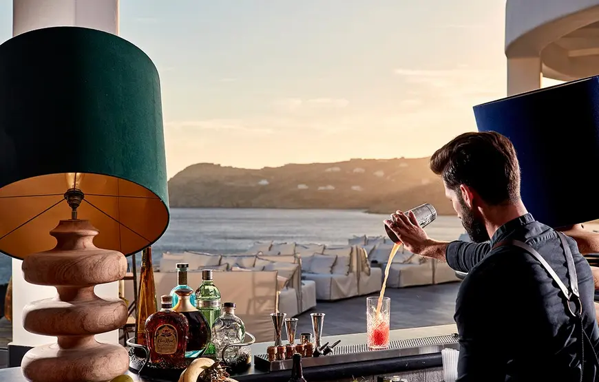 un-barman-sta-servendo-un-bicchiere-alcoolico-arancione-davanti-a-un-mare-argento-con-luce-al-tamonto