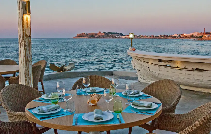 un-tavolo-apparecchiato-sul-mare-una-barca-bianca-vicino-sullo-sfondo-il-mare-e-una-roccia-colorata-di-rosso-al-tramonto
