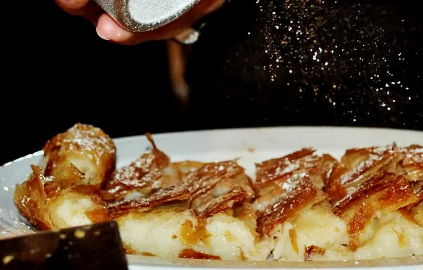 dolce-tipico-greco-una-sfoglia-ripiena-di-crema-con-caramello-sopra-mentre-una-mano-spolvera-dello-zucchero-a-velo-si-chiama-bougatsa