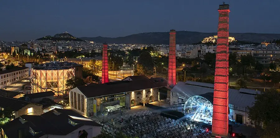 quartiere-di-gazi-atene-con-i-simboli-delle-ciminiere-rosse-e-una-vista-dall-alto-della-piazza-dove-si-sta-svolgendo-un-concerto-la-città-è-illuminata-di-sera