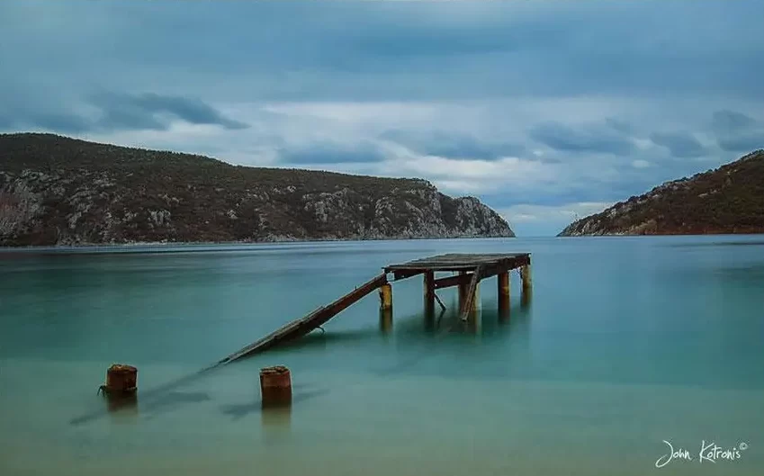un-ponticello-crollato-sulla-riva-una-distesa-di-acqua-del-porto-koufo-e-dietro-delle-montagnole-basse