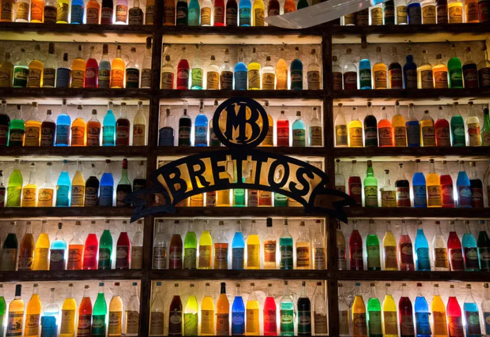 parete-rivestita-da-una-scaffalatura-in-legno-con-bottiglie-di-vetro-colorate-retro-illuminazione-al-centro-c-è-scritto-brettos-in-ferro