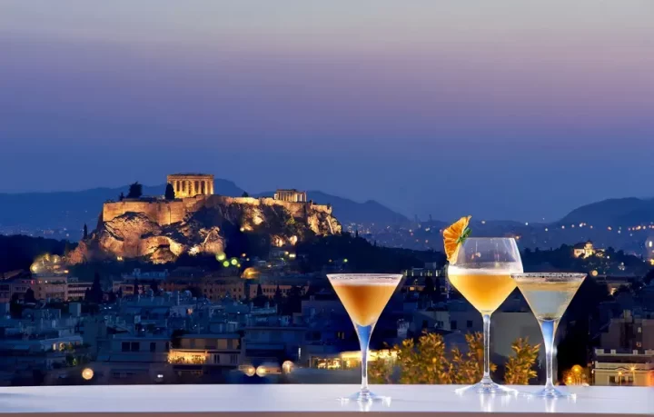 galaxy-bar-hilton-ad-atene-per-un-capodanno-eccezionale due-drink-arancioni-e-bicchieri-ghiacciati-sono-su-una-balaustra-che-guarda-l'acropoli-di-atene-illuminata-sullo-sfondo