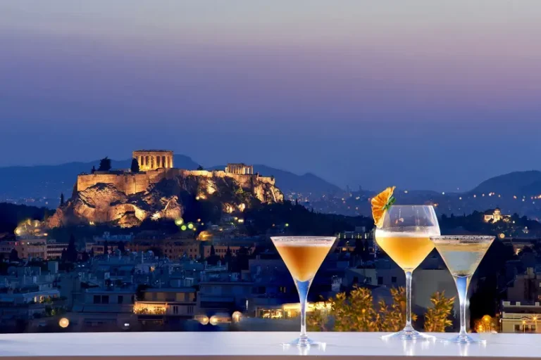 due-drink-arancioni-e-bicchieri-ghiacciati-sono-su-una-balaustra-che-guarda-l'acropoli-di-atene-illuminata-sullo-sfondo