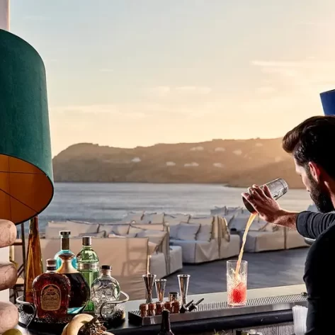 un-barman-sta-servendo-un-bicchiere-alcoolico-arancione-davanti-a-un-mare-argento-con-luce-al-tamonto