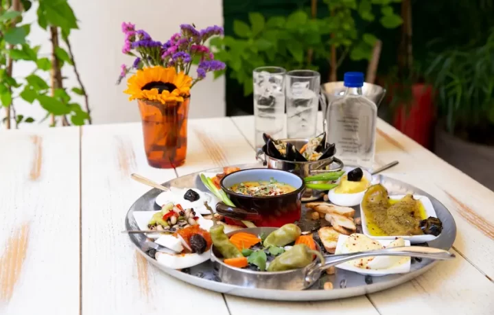 brunch-mezedes-con-ouzo-salonicco tavolino-con-sopra-un-piatto-pieno-di-stuzzichini-tra-olive-e-pezzetti-di-pesce-dietro-due-bicchieri-di-ouzo-e-di-fianco-un-vasetto-con-un-girasole-e-fiorellini-violetti