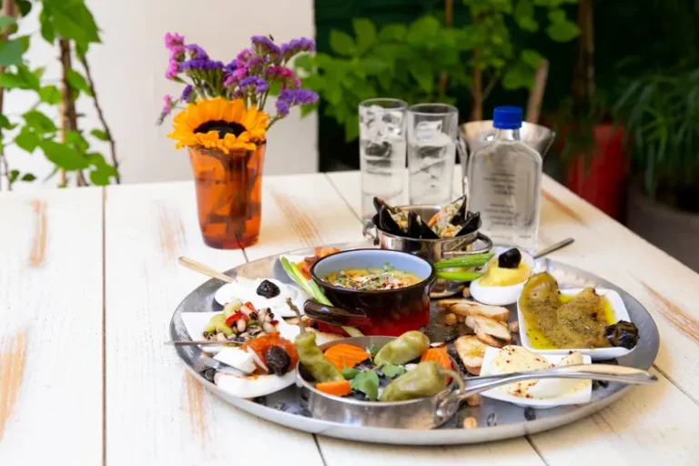 tavolino-con-sopra-un-piatto-pieno-di-stuzzichini-tra-olive-e-pezzetti-di-pesce-dietro-due-bicchieri-di-ouzo-e-di-fianco-un-vasetto-con-un-girasole-e-fiorellini-violetti