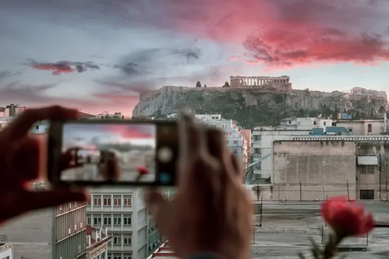 una-mano-solleva-un-cellulare-che-inquadra-l'acropoli-dietro-si-apre-il-paesaggio-ateniese