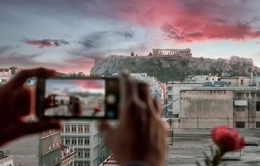 dal-polis-grand-foto-dell-acropoli-vista-da-dietro-un-cellulare-plisgrandhotel.gr una-mano-solleva-un-cellulare-che-inquadra-l'acropoli-dietro-si-apre-il-paesaggio-ateniese