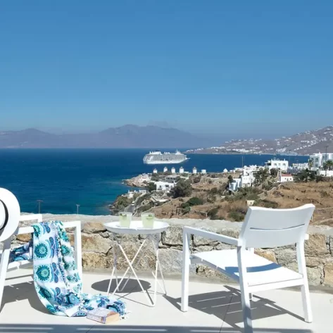 terrazza-con-due-sedie-bianche-un-tavolino-che-guardano-la-grecia-dall-alto-il-mare-sotto-e-le-casette-bianche