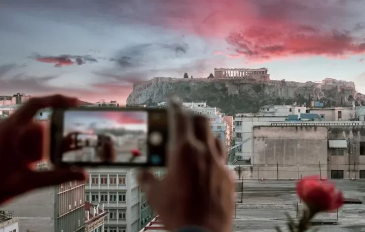 una-mano-solleva-un-cellulare-che-inquadra-l'acropoli-dietro-si-apre-il-paesaggio-ateniese